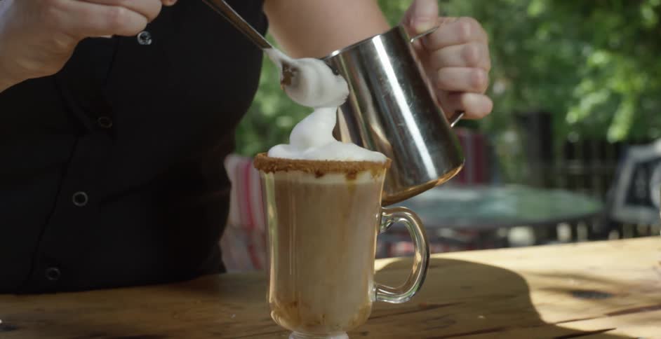 A woman pours cream into a glass of coffee on an outdoor table at midday using a stainless steel cup