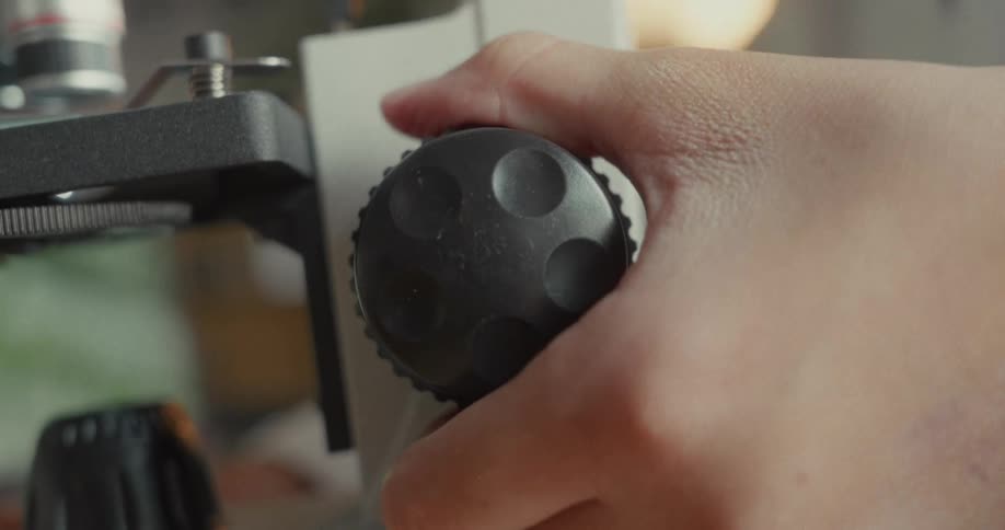 A hand is adjusting the knob on a microscope in a lab