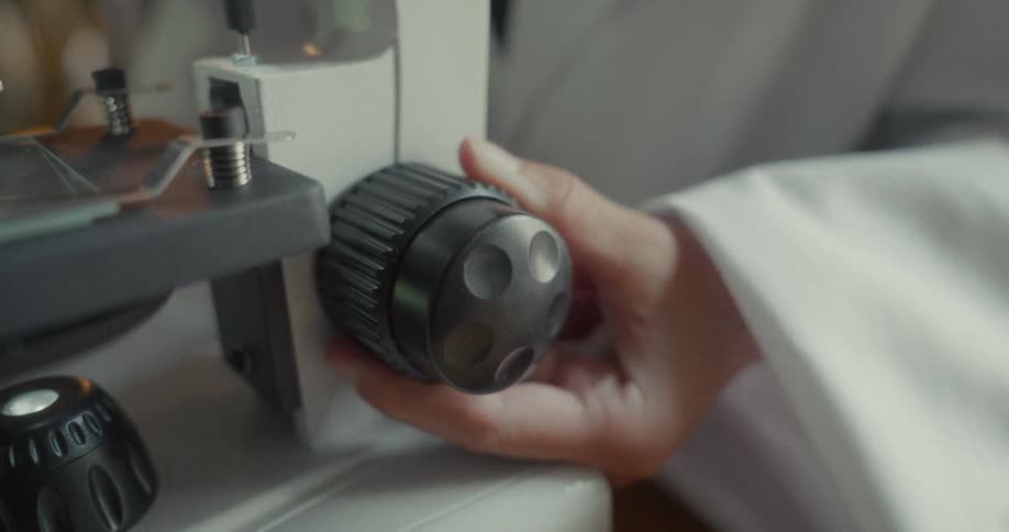 A person wearing a white lab coat is using a microscope and adjusting the knob