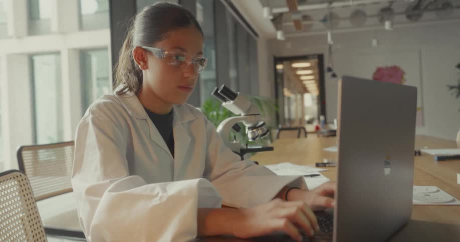 Woman in glasses and a lab coat sitting at a table typing on a laptop
