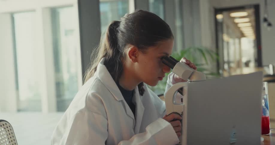 The girl leans in her chair to look into the microscope on the table