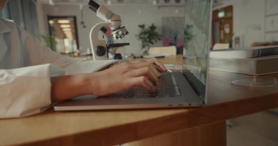 A girl in a lab coat types on a laptop beside a microscope in a bright laboratory