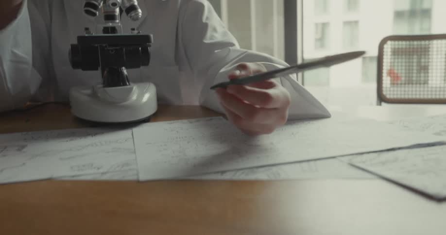 A young female scientist makes notes after viewing a microscope slide at a table in a lab during daytime