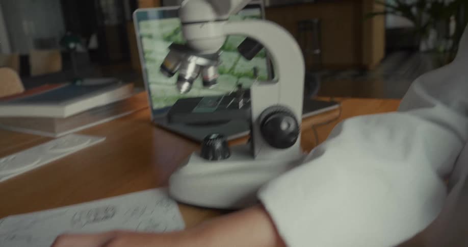 A woman in a lab coat circles a sketch with a red marker on a sheet of drawings while working beside a microscope and a laptop in a laboratory filled with scientific tools
