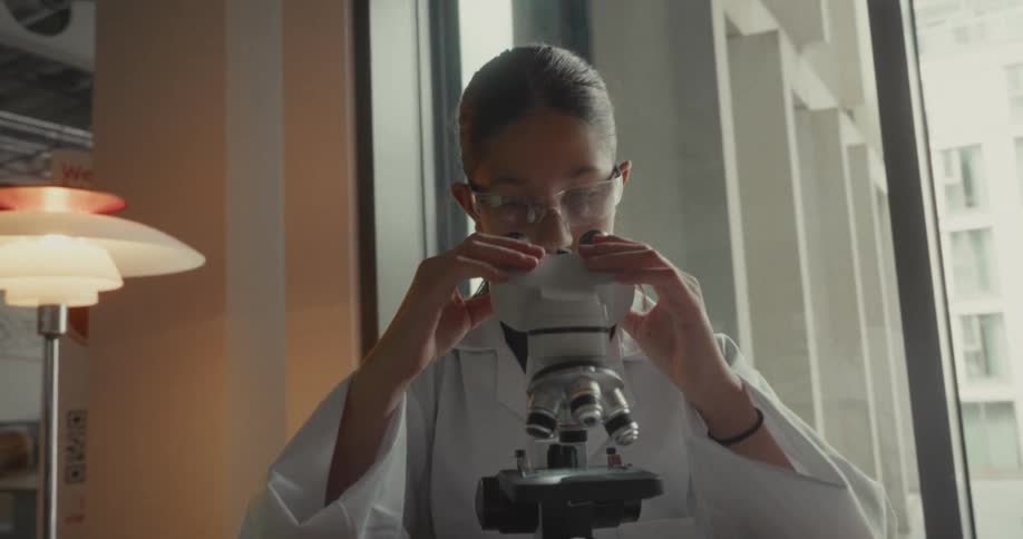 Young female scientist notes findings while viewing a slide on a microscope in a lab during daytime