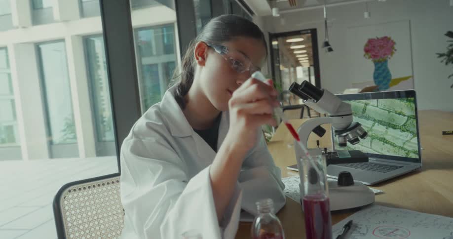 Young female scientist collects a red liquid sample with a pipette from a bottle and drops it into a cylindrical beaker on a desk in a lab during daytime