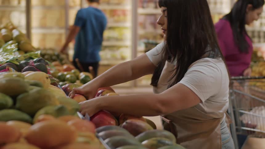 The video shows a woman with dark hair and bangs wearing a white tshirt and an apron meticulously arranging a display of mangoes in a grocery store she carefully places each mango on the shelf ensuring a neat presentation in the background