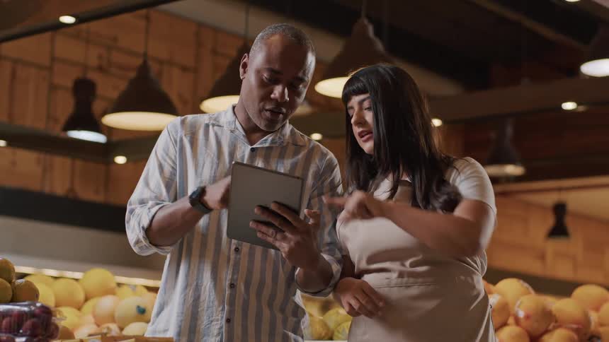 A female store attendant is assisting a customer as they go through a tablet together looking at produce while standing in the fruit section of the store