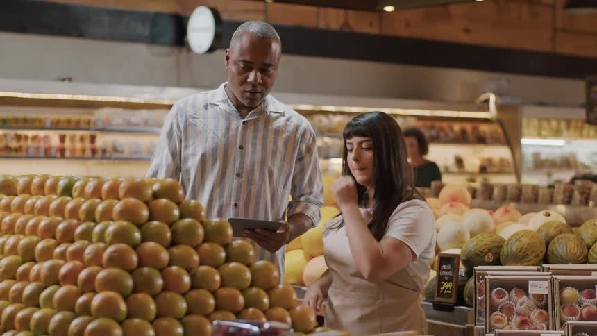 The video features two individuals a man and a woman standing next to a large display of oranges in what appears to be a grocery store the man is holding a tablet and seems to be discussing something with the woman who is wearing an apron and engaged