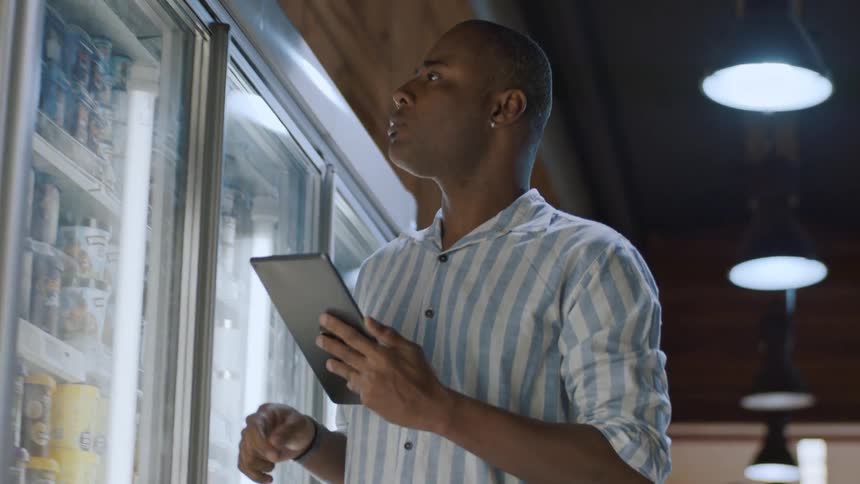 A man in a striped shirt holding a tablet and looking at a refrigerated display case in what appears to be a store he seems to be checking inventory or prices as he looks from the tablet to the items in the cooler