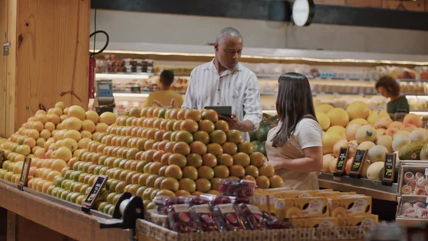 A manager checks a tablet while explaining the fresh produce section to an attentive employee in a grocery store