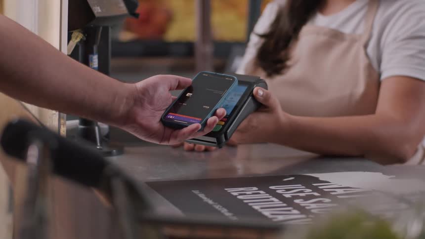 Closeup of a contactless payment transaction a person is holding a smartphone near a payment terminal indicating they are making a payment a store employee identifiable by their apron is holding the payment terminal
