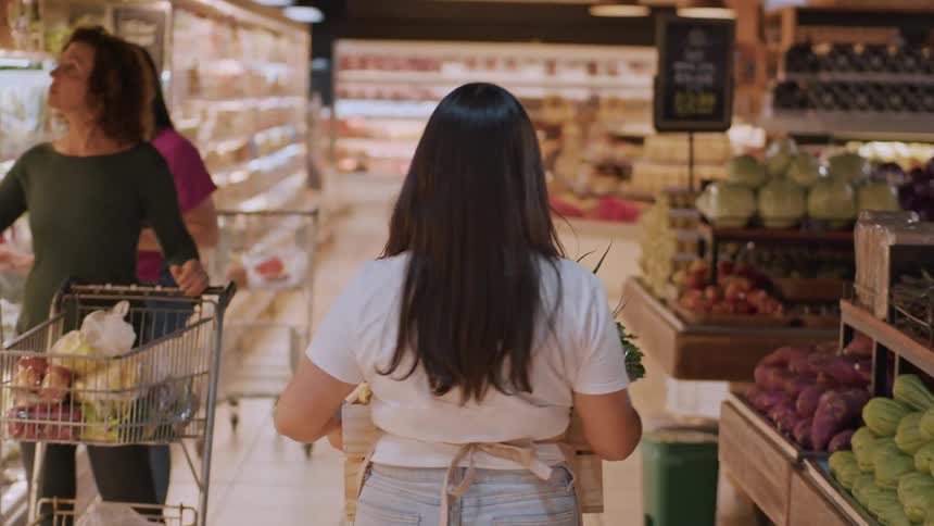 Grocery store clerk carries a box through the produce section passing multiple customers