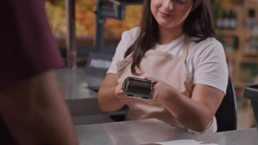 A man in a store ready to pay for his items hands his credit card to the female cashier she inserts it into the pos machine and hands it to him to enter his pin and complete the payment