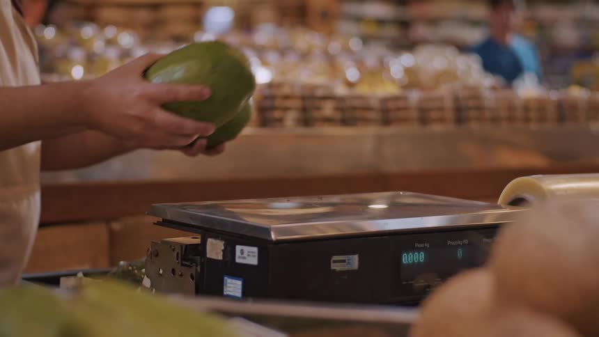 A person sets two large avocados on a digital scale at a market counter