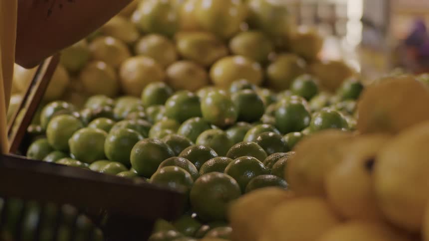 A hand selects limes from a mixed pile of lemons and limes at a market stall