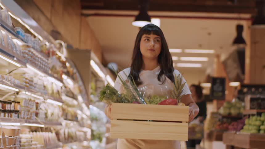 An employee carries a wooden crate filled with assorted fresh vegetables through a grocery store aisle lined with produce
