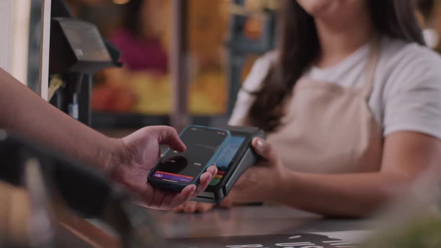 A closeup of a contactless payment transaction a person is holding a smartphone near a payment terminal indicating they are making a payment a store employee identifiable by their apron is holding the payment terminal