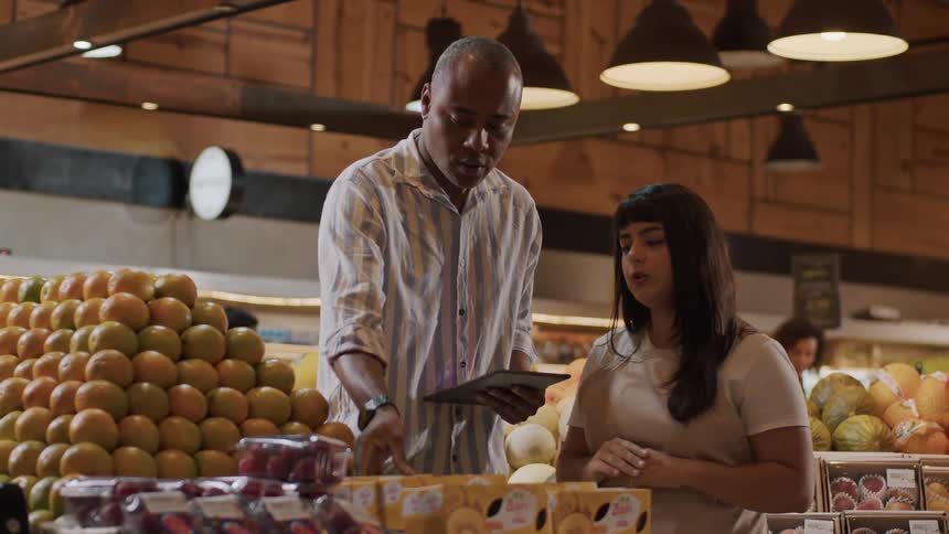 A manager checks a tablet while explaining the fresh produce section to an attentive employee in a grocery store