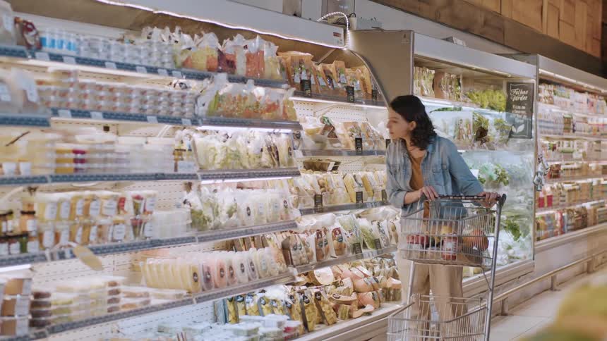 A woman pushes a shopping cart past a refrigerated section stocked with assorted packaged goods in a brightly lit grocery store