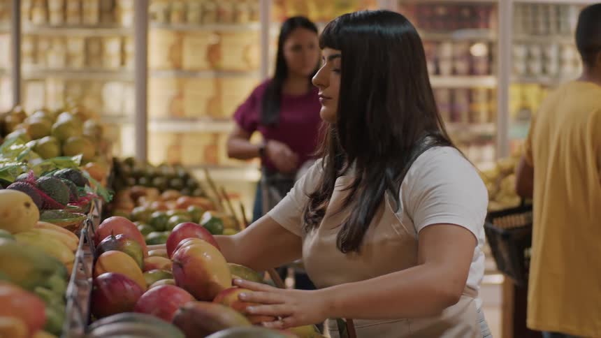 A woman with dark hair and bangs wearing a white tshirt and an apron arranging mangoes on a display shelf in a grocery store she carefully picks up and places the mangoes ensuring they are neatly presented in the background