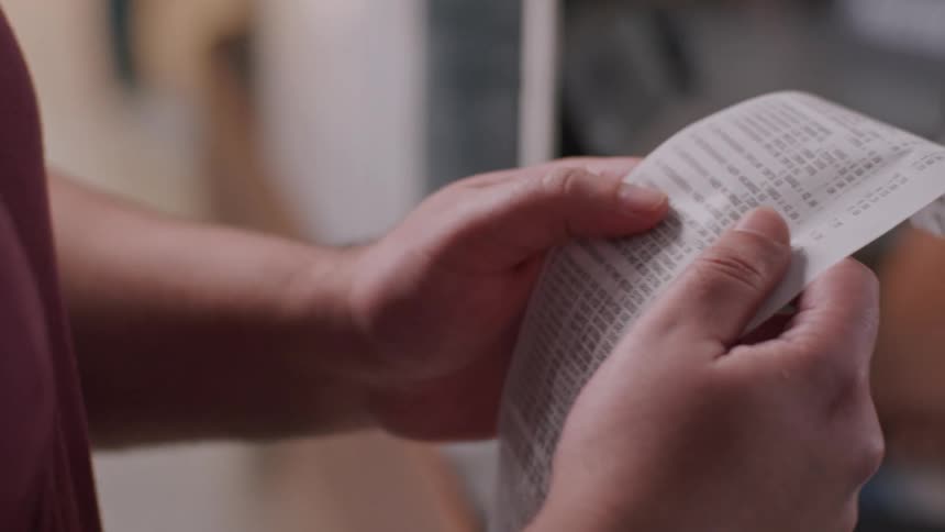 A closeup of a persons hands holding and examining a long paper receipt the persons fingers are moving appearing to unfold or review the details printed on the receipt