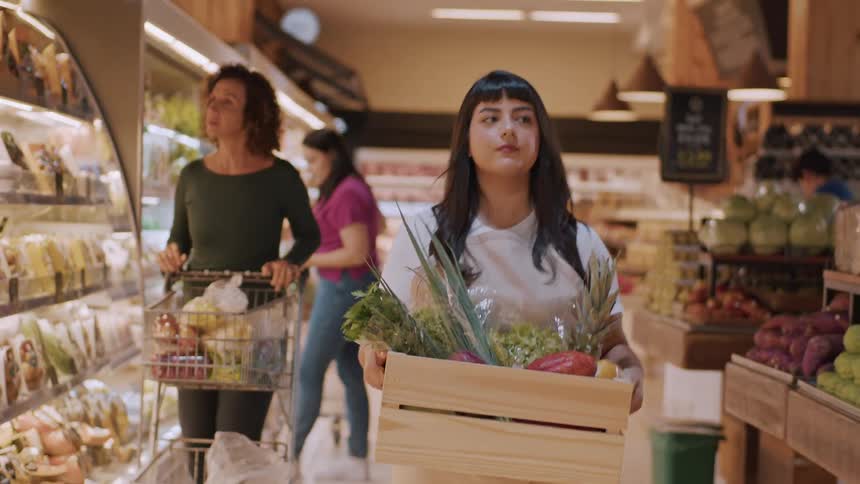 Woman walks through a supermarket holding a wooden box full of fresh fruits and vegetables