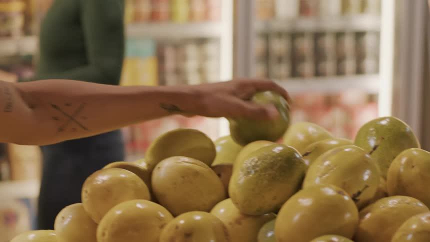 The video shows a closeup of a persons hand sorting through a pile of passion fruits in a grocery store the hand picks up several fruits inspects them and places them back likely checking for ripeness or quality in the background