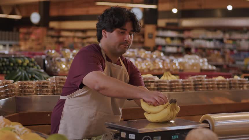 A female customer hands a bunch of bananas to a market employee who weighs them and returns them with a smile