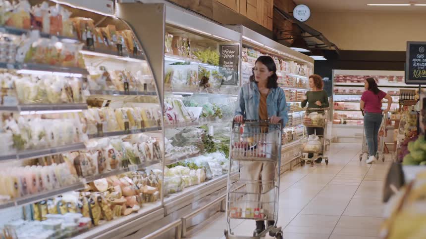 A woman pushes a shopping cart through a grocery store aisle browsing the refrigerated products
