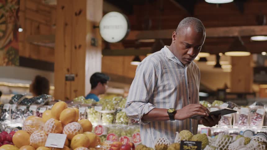 A person checks a tablet near stacked fruit boxes in a brightly lit grocery store produce section