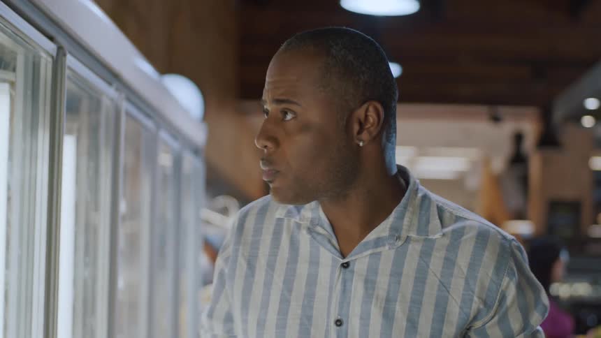 A man in a striped shirt standing in front of a refrigerated display case likely in a supermarket he is looking intently at the items inside the cooler seemingly deciding what to purchase