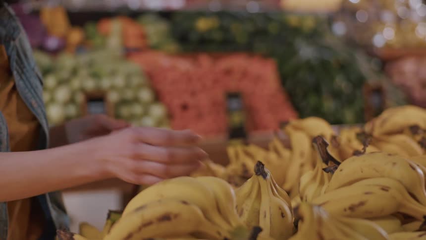 The video shows a woman in a denim jacket and a yellow shirt selecting a bunch of ripe yellow bananas from a display in a grocery store she carefully examines the bananas before deciding to take them