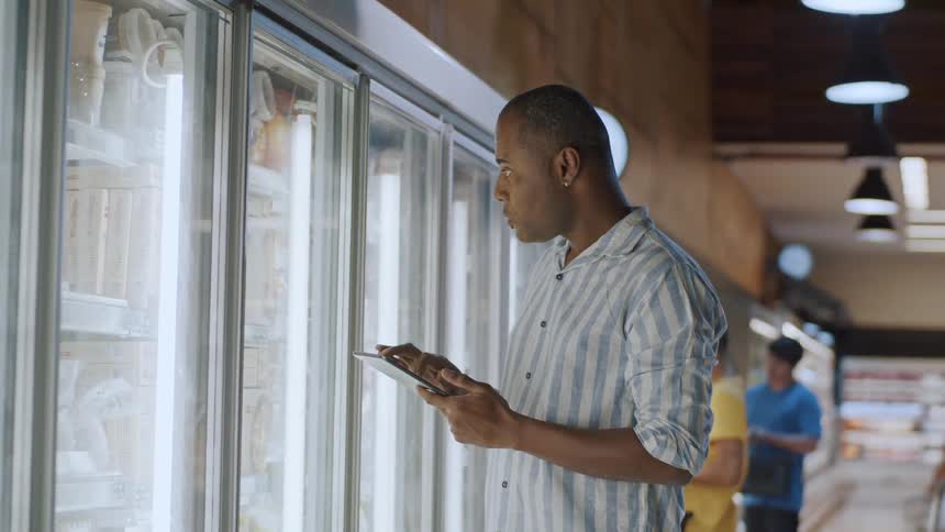 A grocery store worker checks inventory in a freezer aisle using a tablet