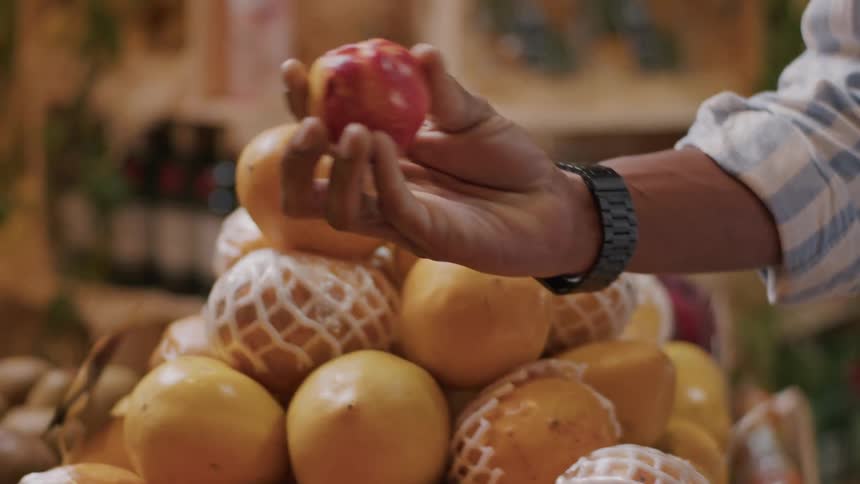 The video shows a closeup of a persons hand sorting through various fruits in a grocery store the hand adorned with a watch picks up and examines different fruits including what appear to be plums mangoes some wrapped in mesh and cherries