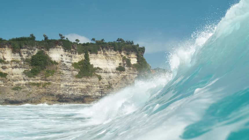 A large ocean wave crashes near a rugged cliffside with sparse vegetation under a clear blue sky