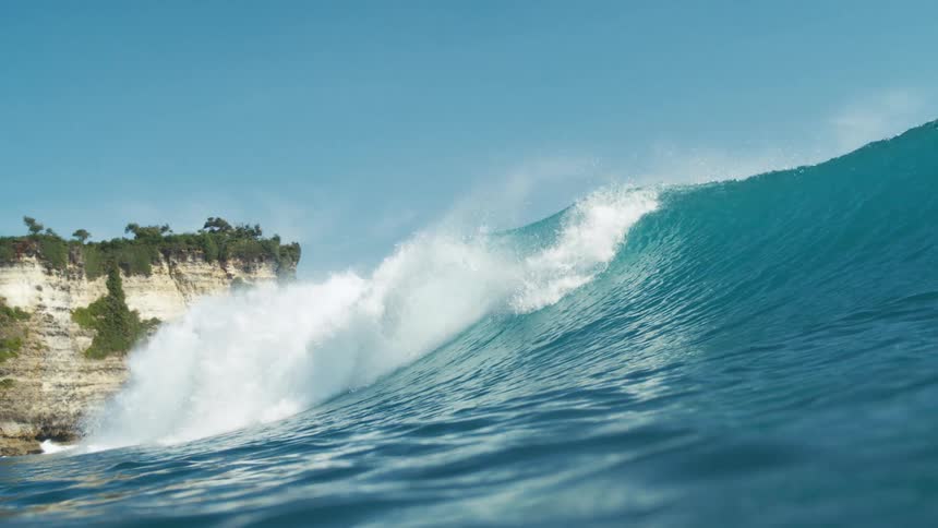 The video displays a vibrant blue ocean with a large wave breaking in the background a lush green cliff face with rocky outcroppings rises against a clear blue sky suggesting a beautiful coastal scene