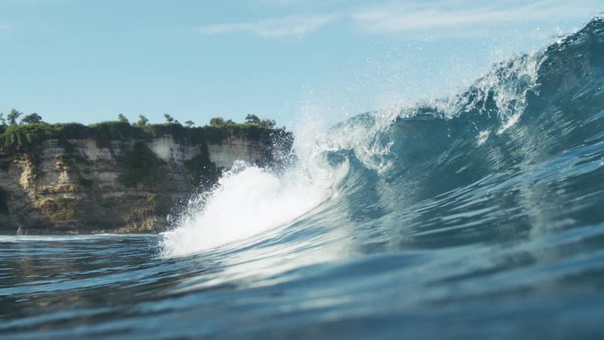 A powerful ocean wave crashes near a rocky coastal cliff under a clear blue sky