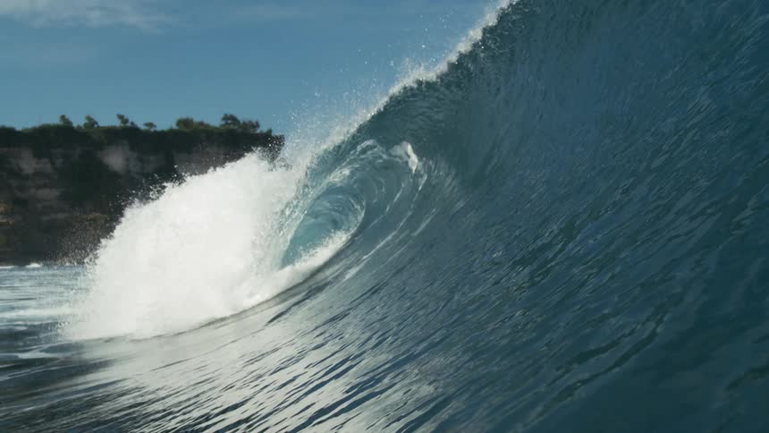 A large ocean wave curls toward a rugged shoreline under a bright blue sky