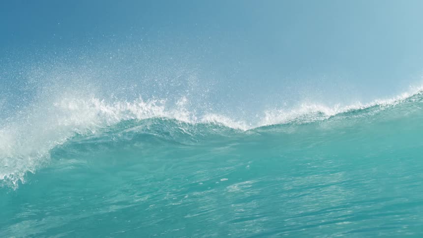 A vibrant blue ocean with a large wave cresting and moving from left to right across the frame the top of the wave is covered in white foam and spray indicating its breaking motion against a bright blue sky with a few faint clouds