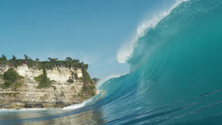 A large ocean wave barrels near a rocky tree covered coastal cliff