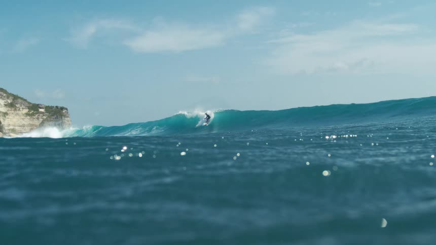 A dynamic underwater shot of a surfer riding a wave the blue ocean water fills the foreground with a large wave visible in the middle ground and a rocky cliff face under a light blue sky in the background the surfer is seen successfully riding waves