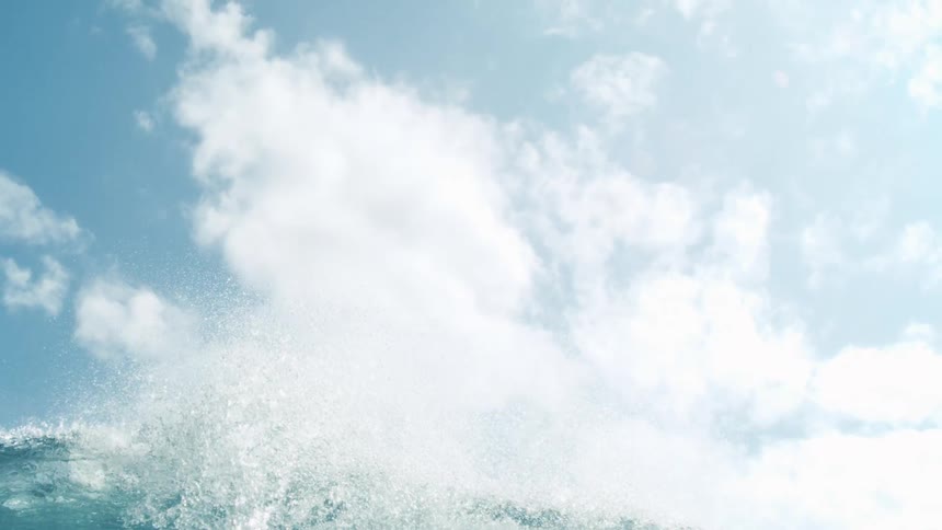 A vibrant blue ocean with a large wave cresting and breaking creating a white foamy splash the clear blue sky above is adorned with scattered white clouds suggesting a bright and sunny day at sea