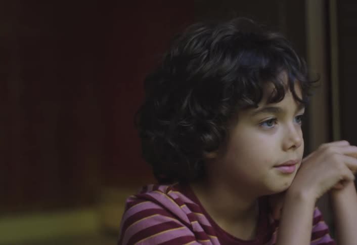A young boy with curly hair looks off to the side with his hands clasped in front of him