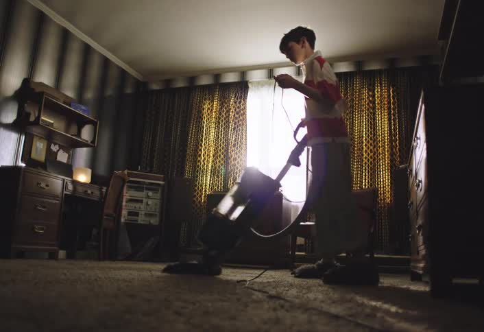 A boy with short dark hair and wearing a red and white striped shirt is seen vacuuming a carpeted room sunlight streams in from a large window with patterned curtains illuminating the dust motes in the air as he cleans