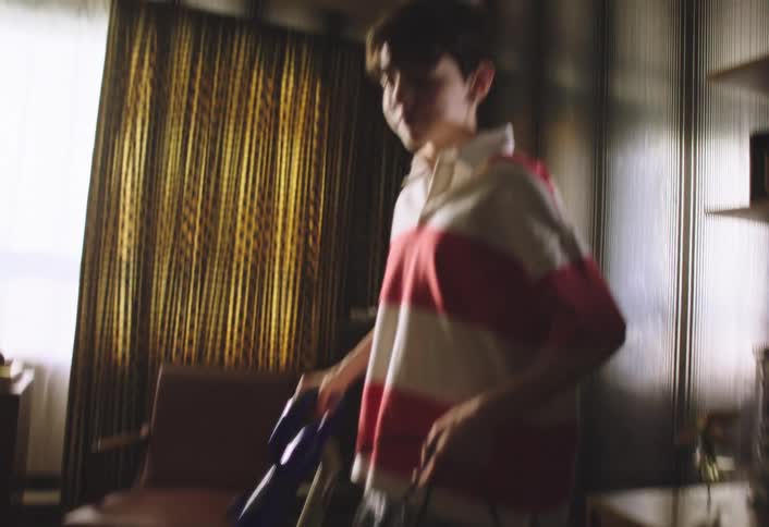 A young person with dark hair wearing a red and white striped polo shirt in what appears to be a bedroom they are operating a blue and gray vacuum cleaner actively moving it across the carpeted floor suggesting they are in the process of cleaning