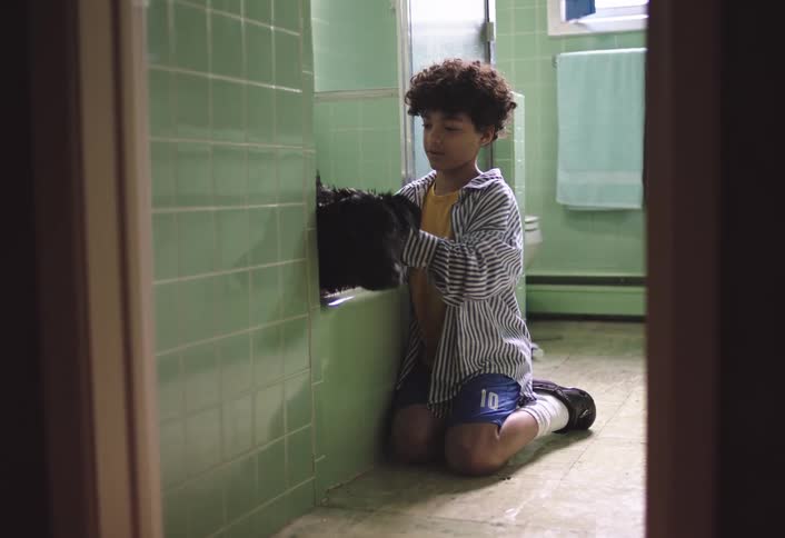 Boy kneels on the bathroom floor while bathing his dog in the bathtub
