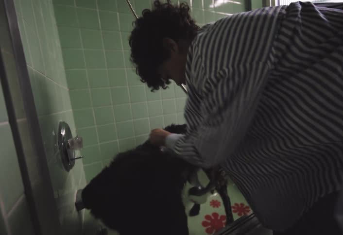 A person wearing a striped shirt and blue shorts is bending over a bathtub attending to a black dog