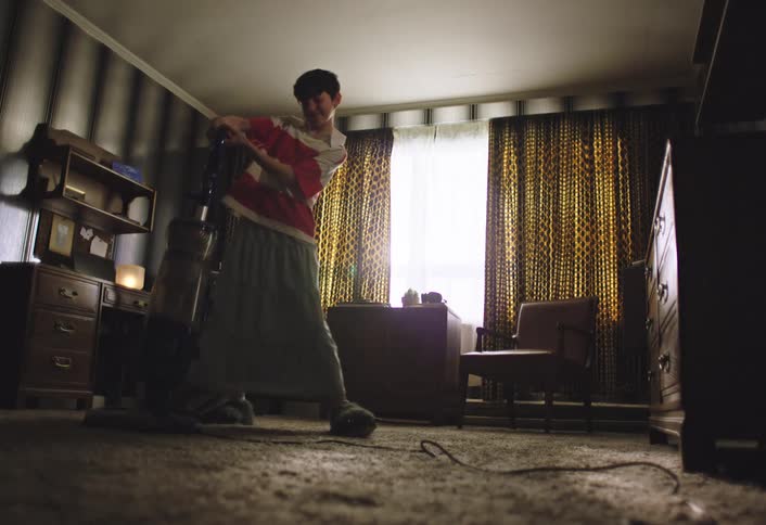 The video shows a child wearing a red and white striped polo shirt and loosefitting pants holding the handle of a vacuum cleaner they are in a room with patterned curtains and wooden furniture and appear to be focused on vacuuming the carpeted floor