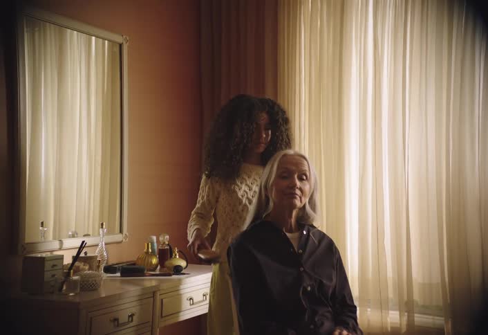 An indoor scene where a person with long curly hair is seen brushing the long straight gray hair of an older woman the older woman is sitting with her eyes closed appearing relaxed while the other person gently combs through her hair